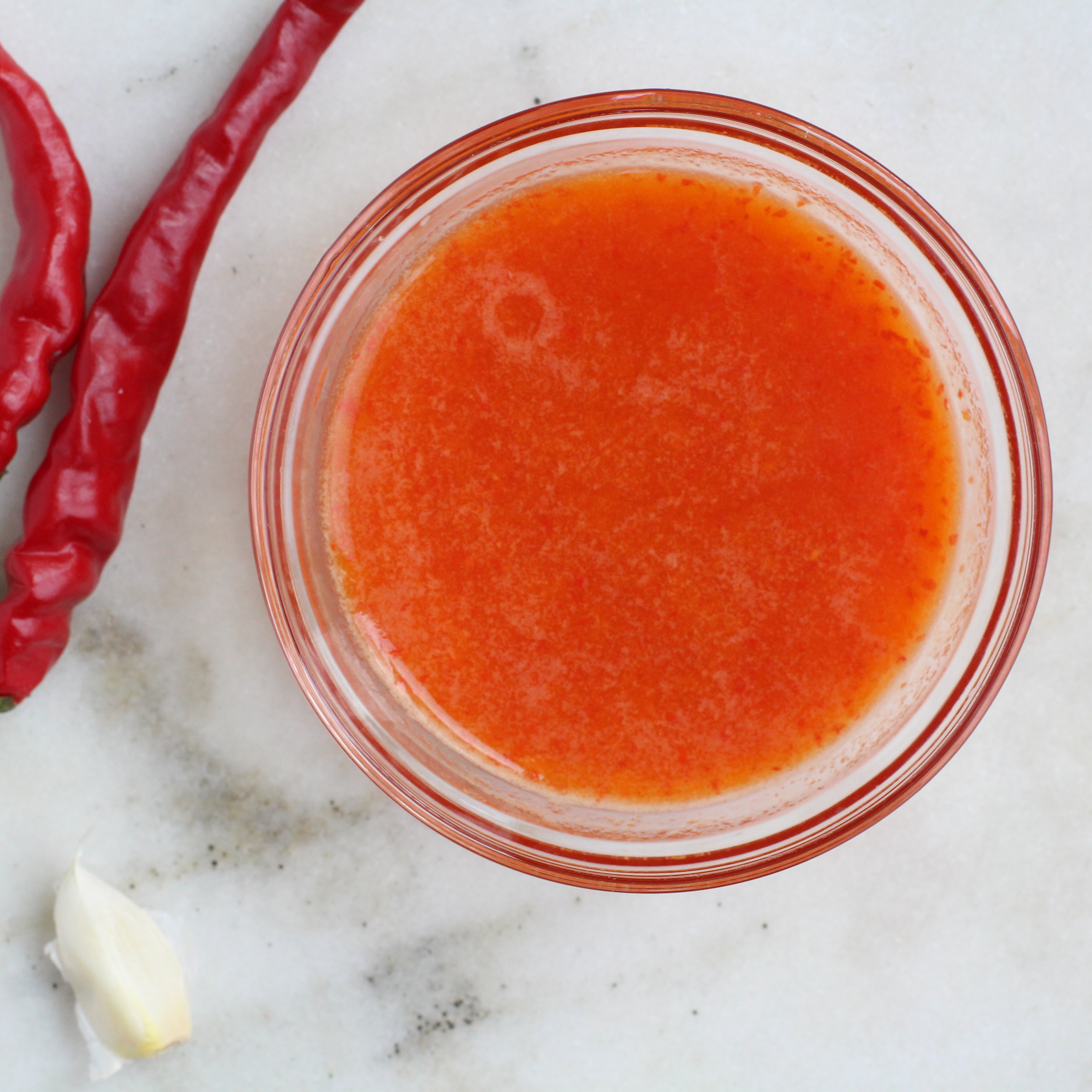 small bowl of healthy hot sauce on a surface with two chilis and a clove of garlic